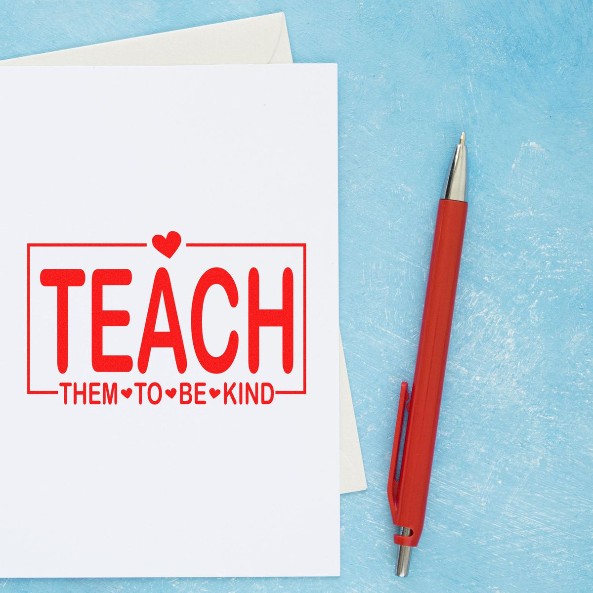 Red teacher rubber stamp reading Teach Them to Be Kind on card, ideal for classroom encouragement and kindness notes.