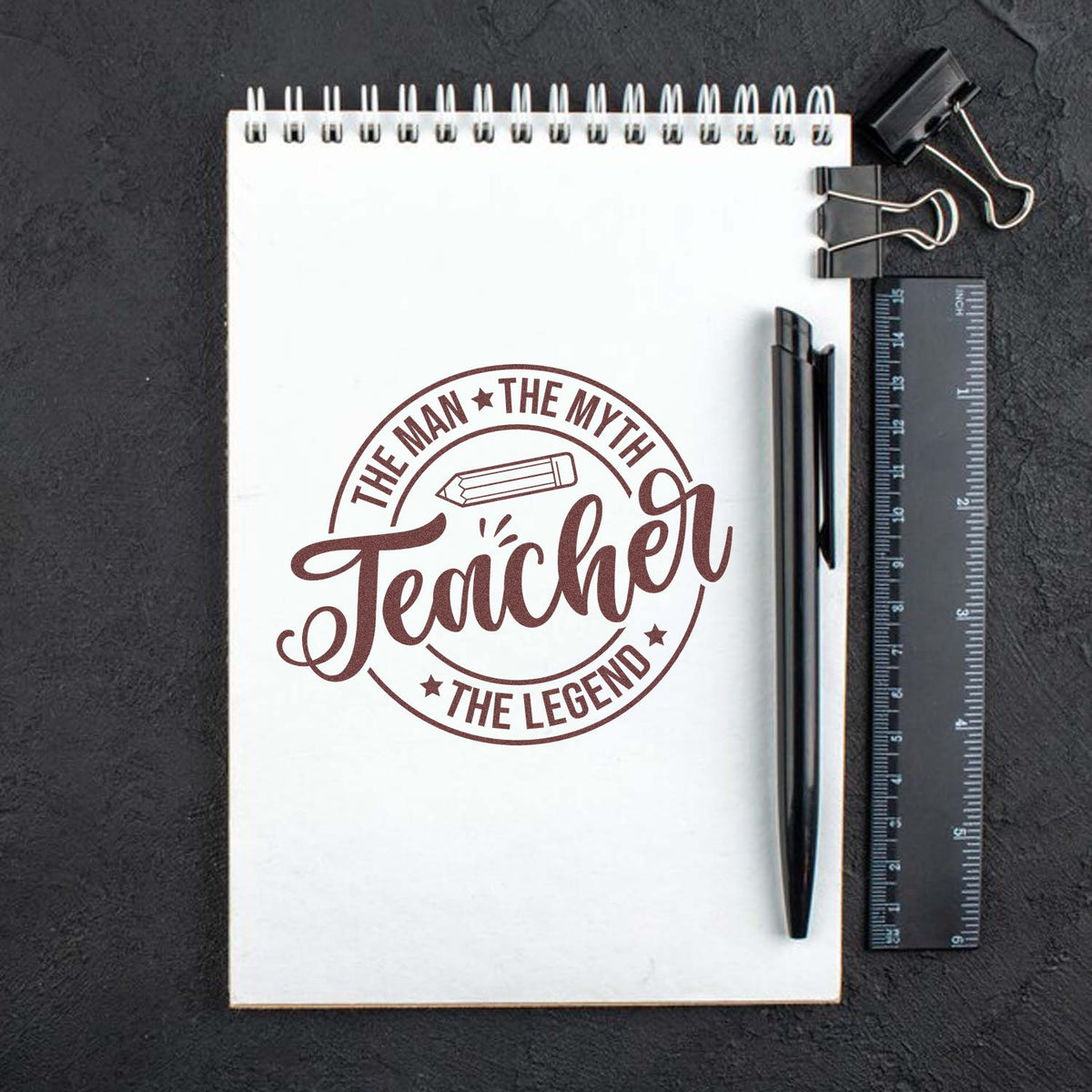 Teacher rubber stamp design reading The Man, The Myth, Teacher, The Legend on a notebook with pen and ruler.