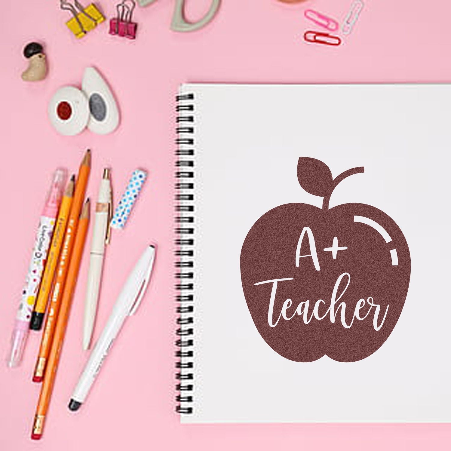 A+ Teacher apple rubber stamp design on notebook, surrounded by pens on pink desk for classroom and planner use.