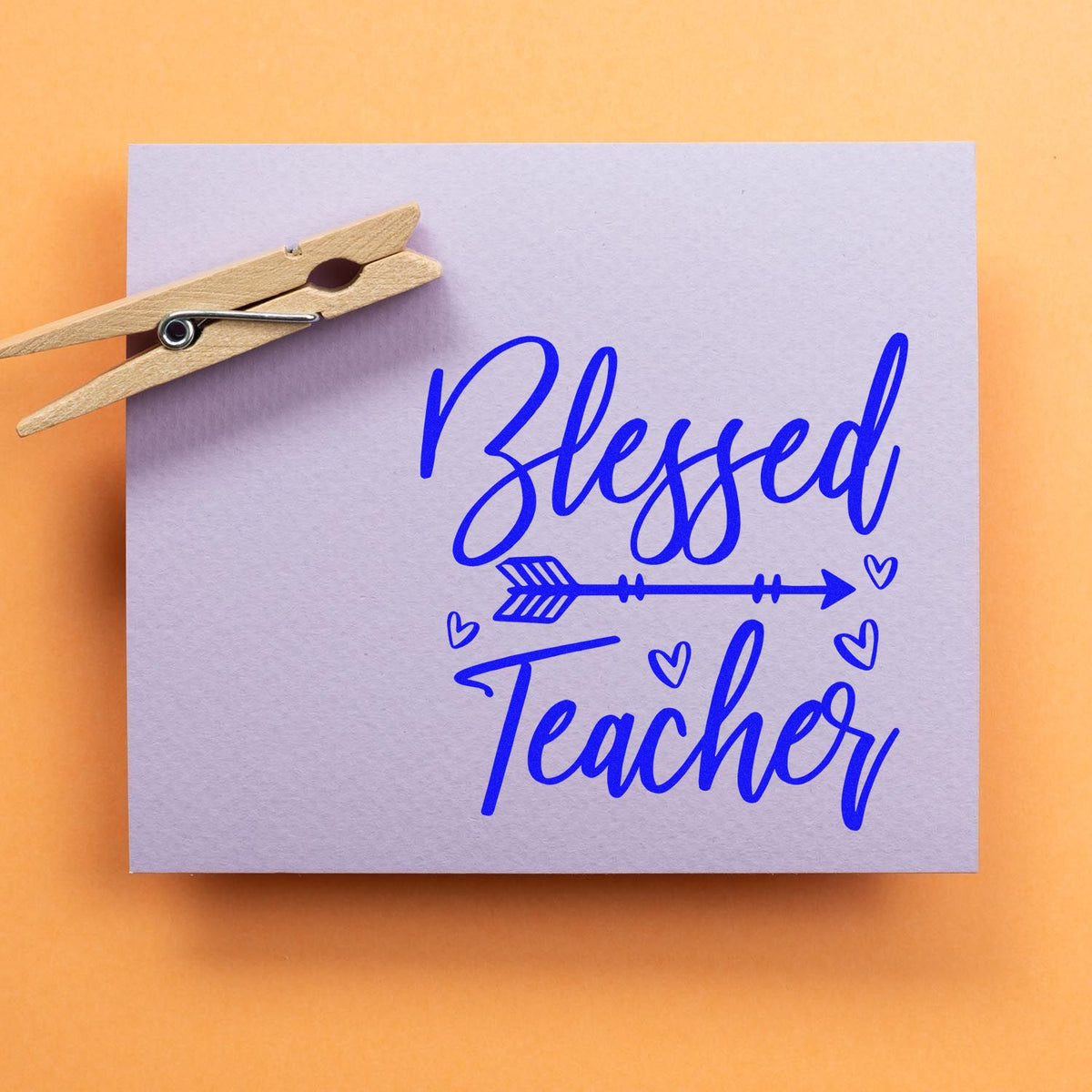 Blessed Teacher rubber stamp design in blue script with arrow and hearts on lavender paper, clipped with a wooden clothespin.