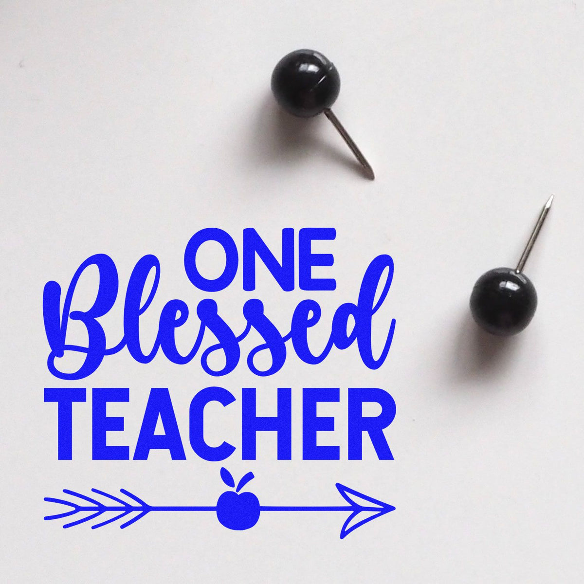 One Blessed Teacher rubber stamp design with blue arrow and apple, shown with push pins on a white background.