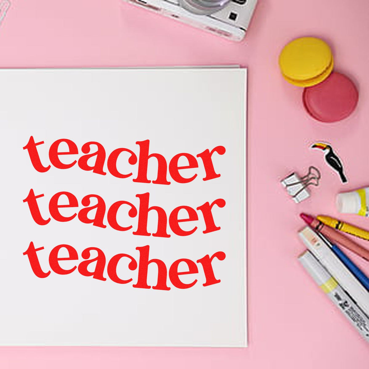 Teacher rubber stamp design stamped three times on paper with craft supplies on a pink background.