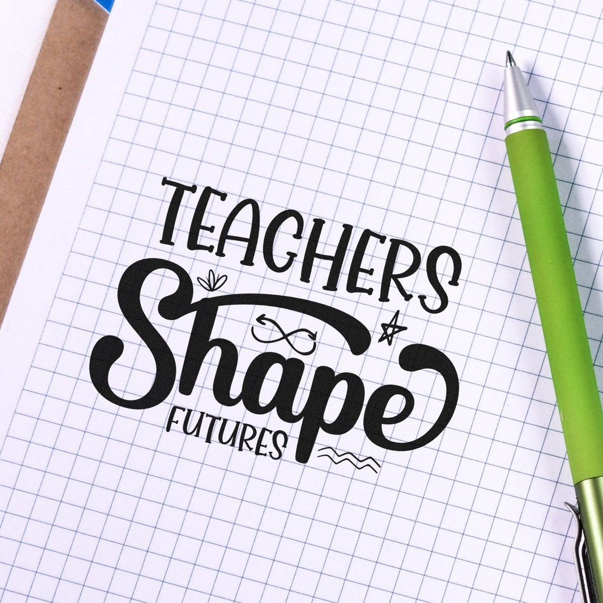 Teacher rubber stamp with Teachers Shape Futures on graph paper beside a green pen, ideal for classroom crafting.