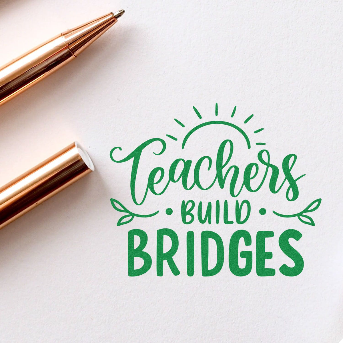 Teacher rubber stamp design reading Teachers Build Bridges with sunburst graphic on white paper beside rose gold pens.