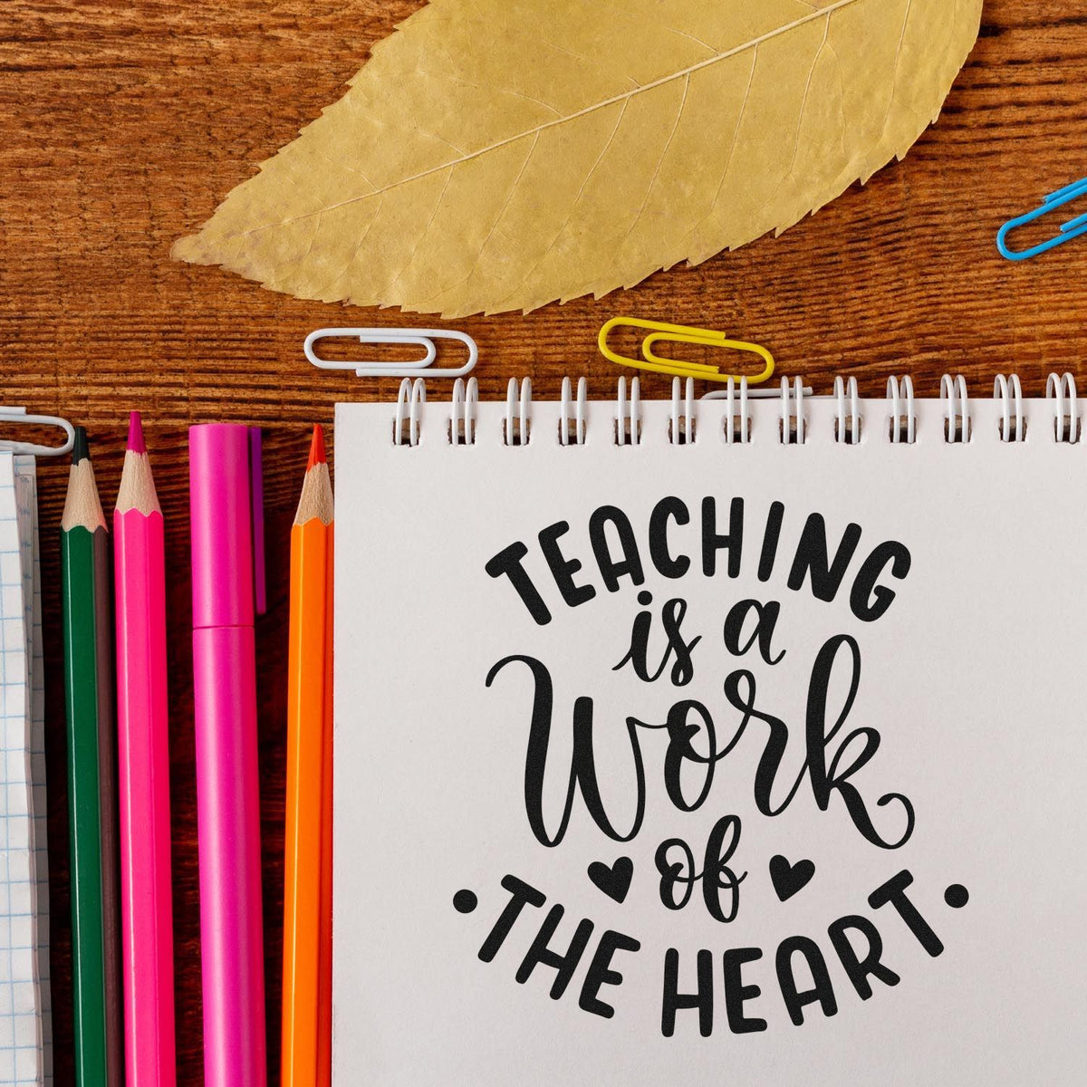 Teacher rubber stamp design reading Teaching is a Work of the Heart on a notebook with pencils and paper clips.
