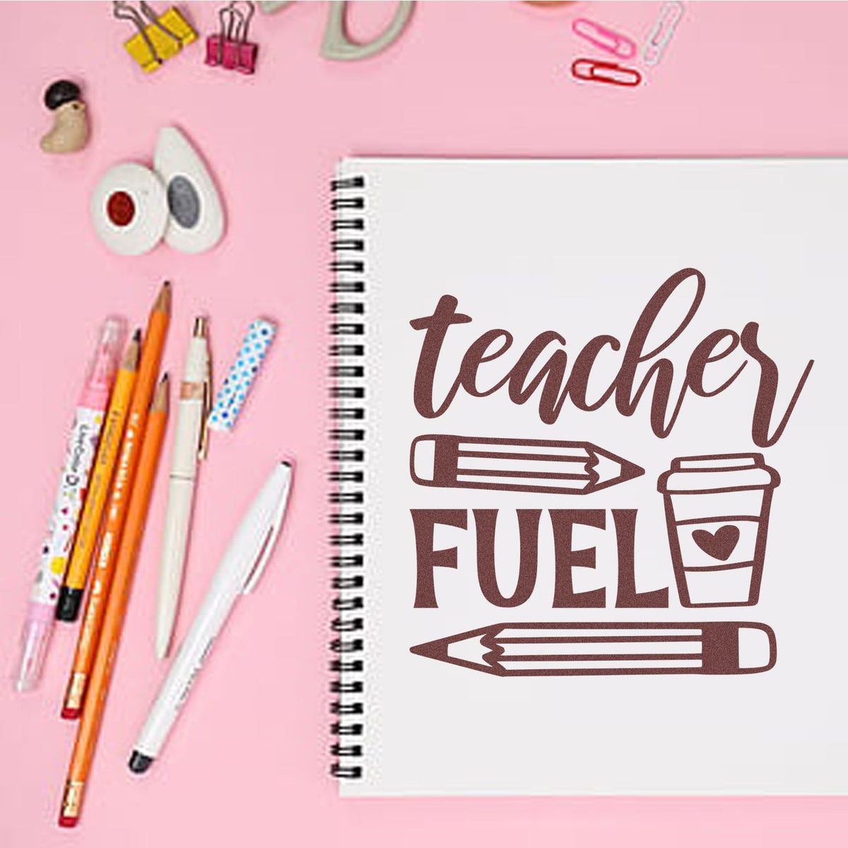 Teacher Fuel rubber stamp design on a spiral notebook, with pencil and coffee cup, surrounded by school supplies on a pink desk.
