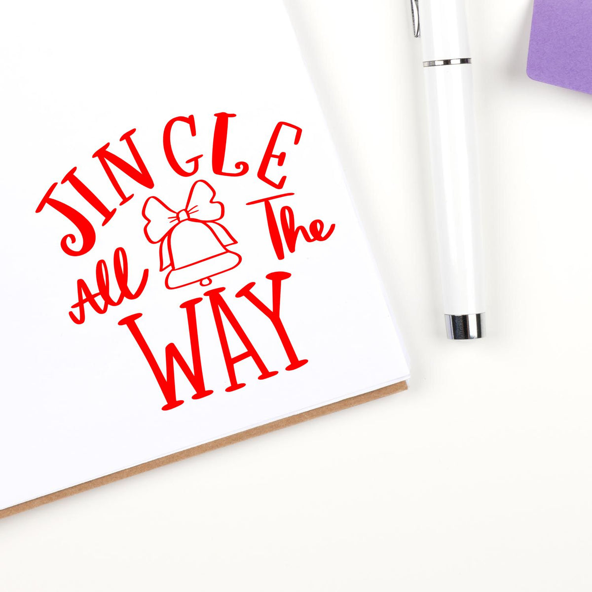 Jingle All the Way Christmas rubber stamp impression in red, with bell icon on paper beside a white pen.