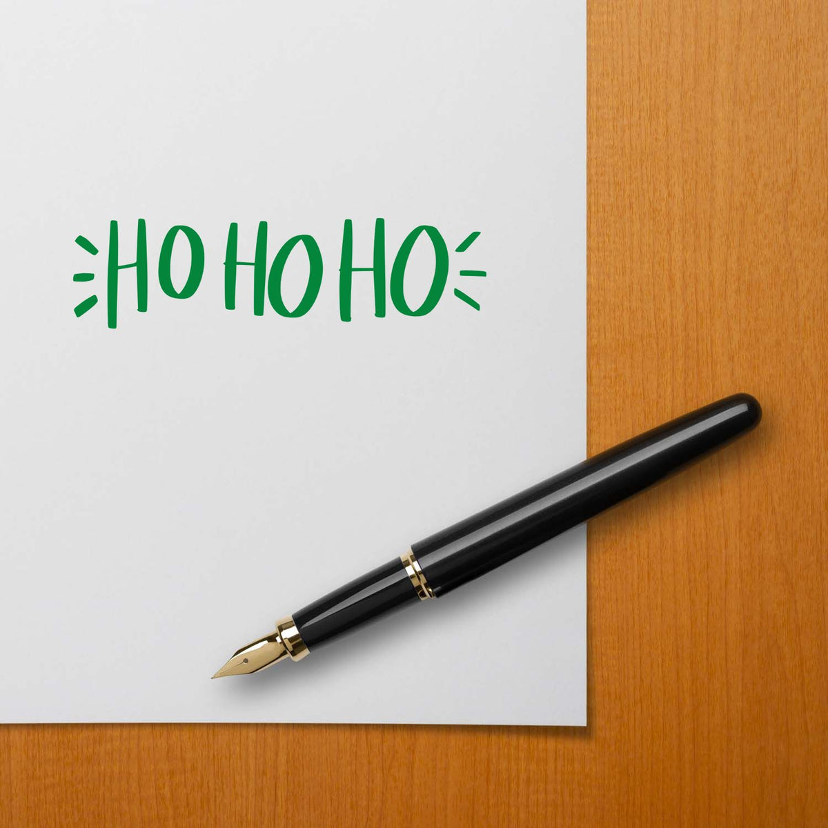 Green HO HO HO Christmas rubber stamp design on paper with fountain pen on a wooden desk.
