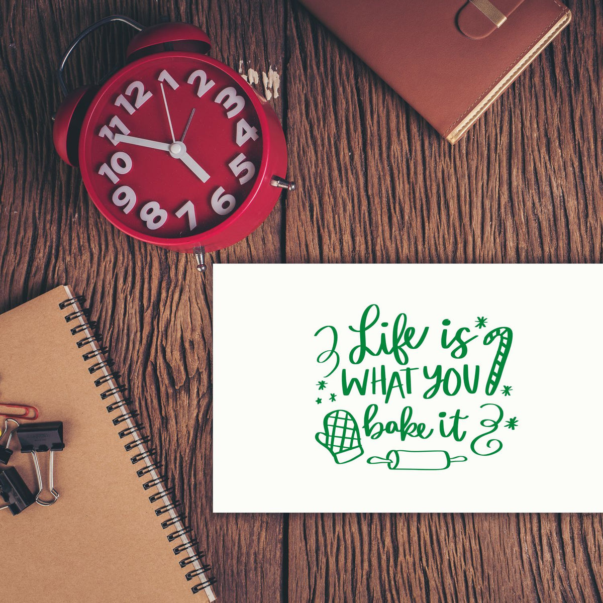 Custom rubber stamp design with baking quote Life is what you bake it, shown on a desk with clock, notebook, and office supplies.