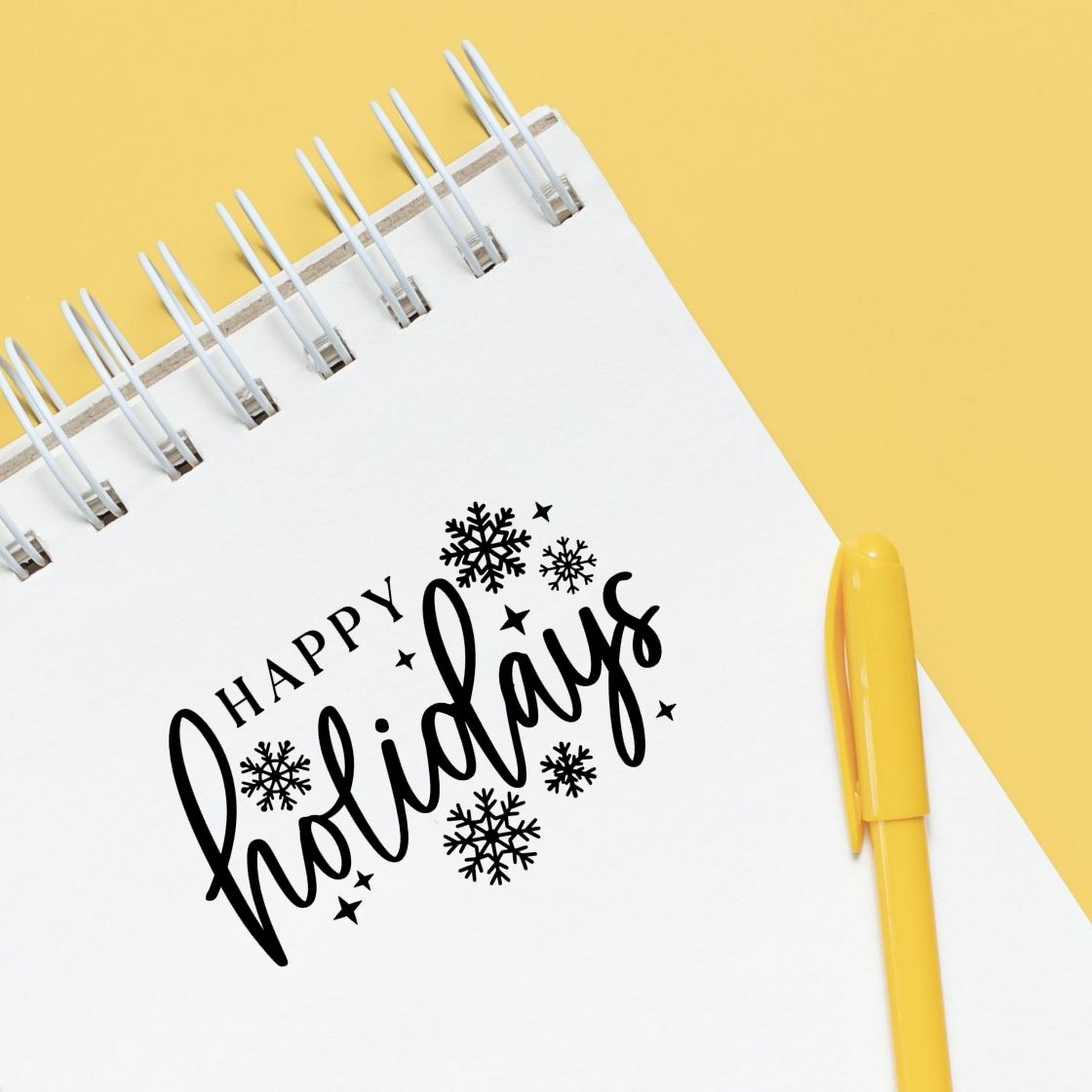 Happy Holidays rubber stamp design with snowflakes on a notebook, styled with a yellow pen.