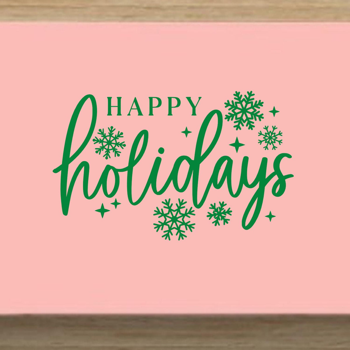 Holiday rubber stamp design with green Happy Holidays lettering and snowflakes on a pink background.