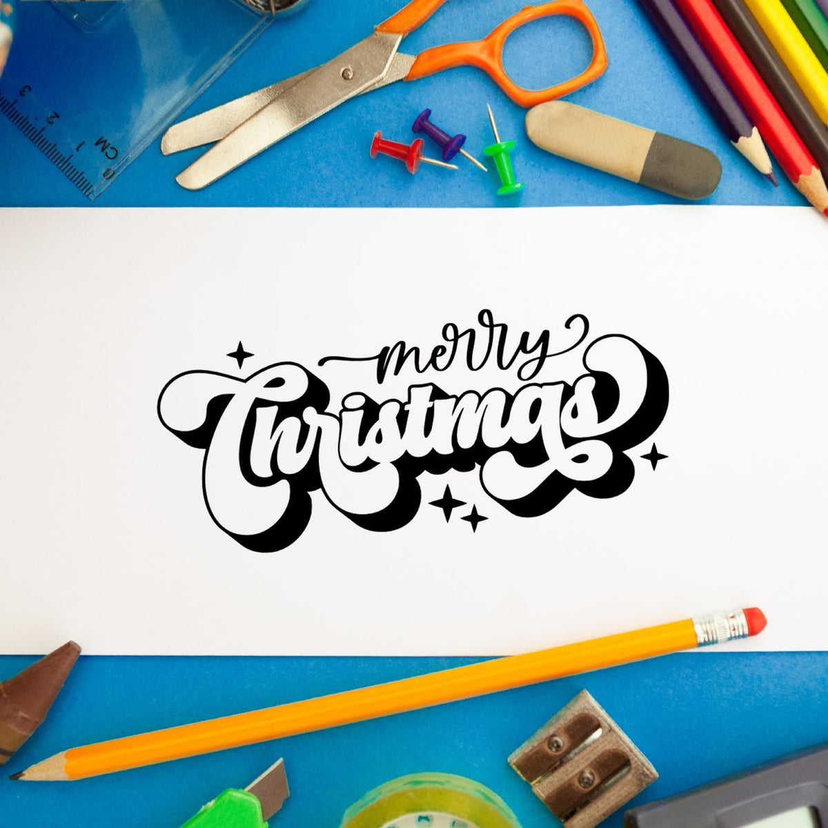 Merry Christmas rubber stamp design on paper surrounded by school supplies for holiday card making and crafts.