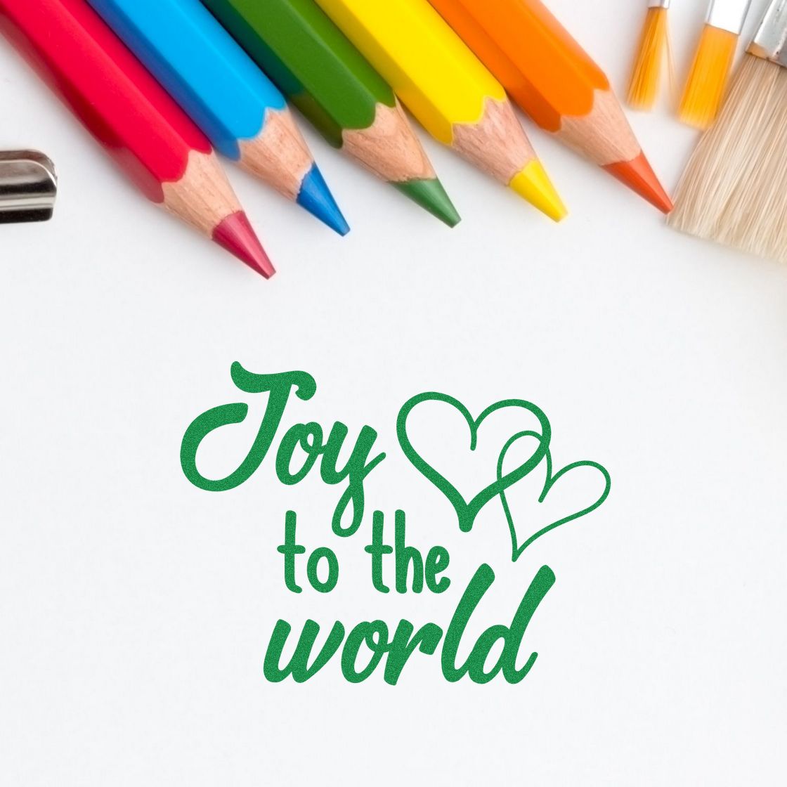 Green Joy to the World rubber stamp design with heart accents, surrounded by colored pencils for holiday card and craft projects.
