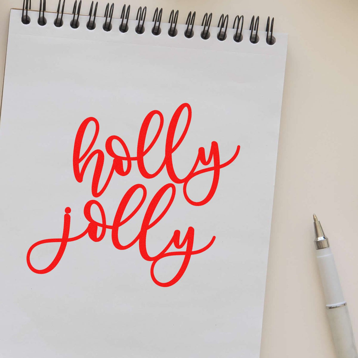 Red holly jolly script rubber stamp design on a spiral notepad with a pen, perfect for festive Christmas crafting.