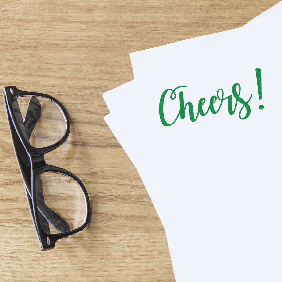 Green Cheers! rubber stamp design on white paper beside eyeglasses on a wooden desk.