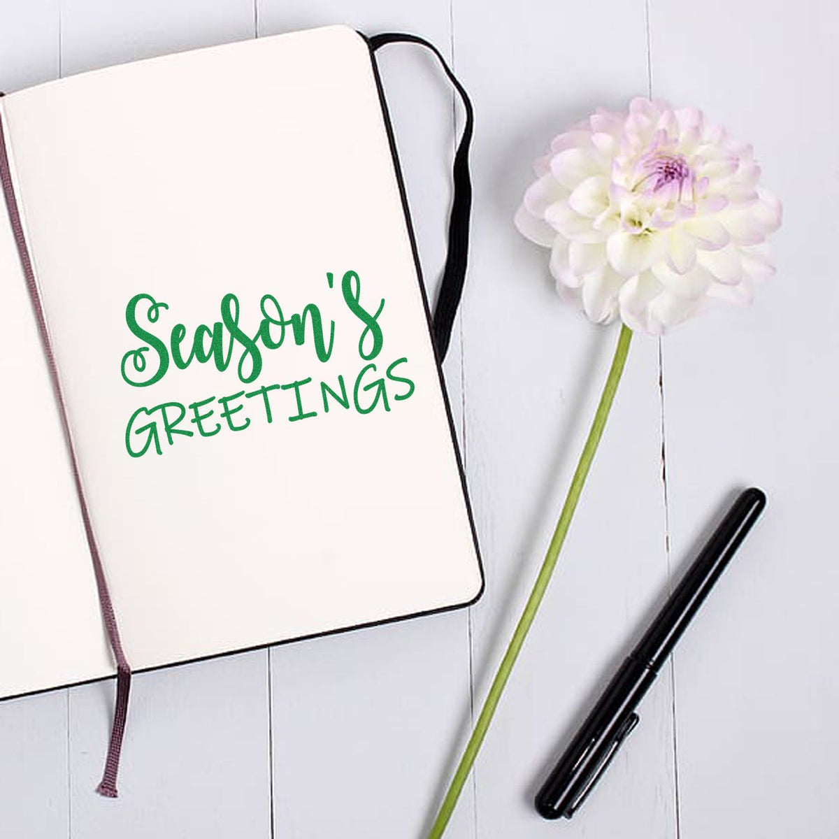 Seasons Greetings rubber stamp design on an open notebook, styled with a flower and pen for holiday card making.