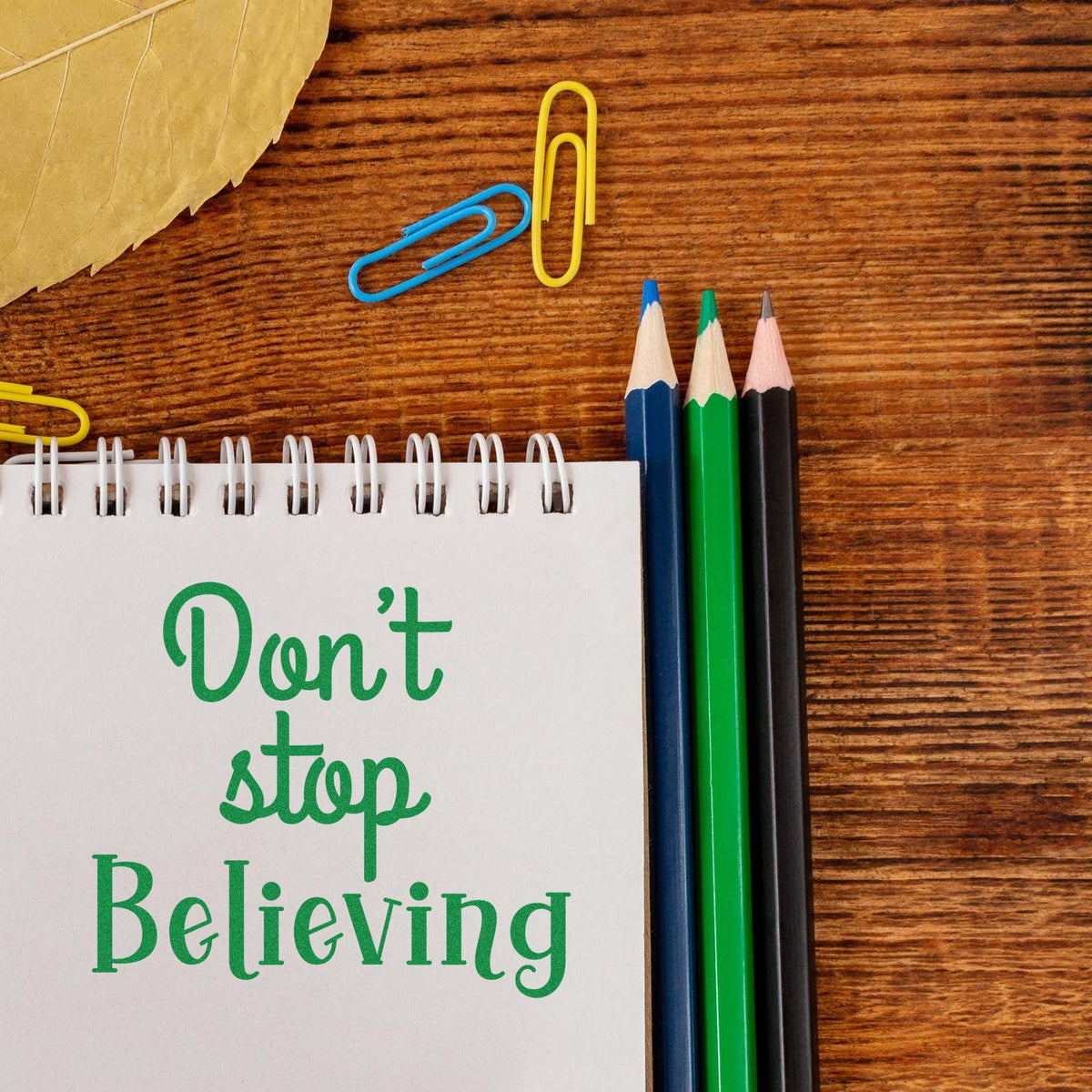 Inspirational Dont stop believing rubber stamp design on a notebook beside pencils and paperclips on a wooden desk.