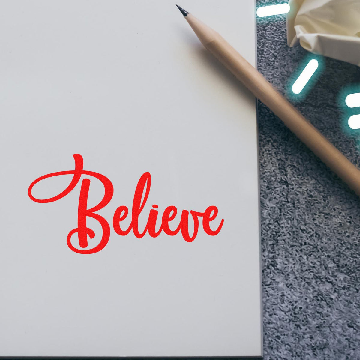 Red Believe script rubber stamp impression on notebook page beside pencil.