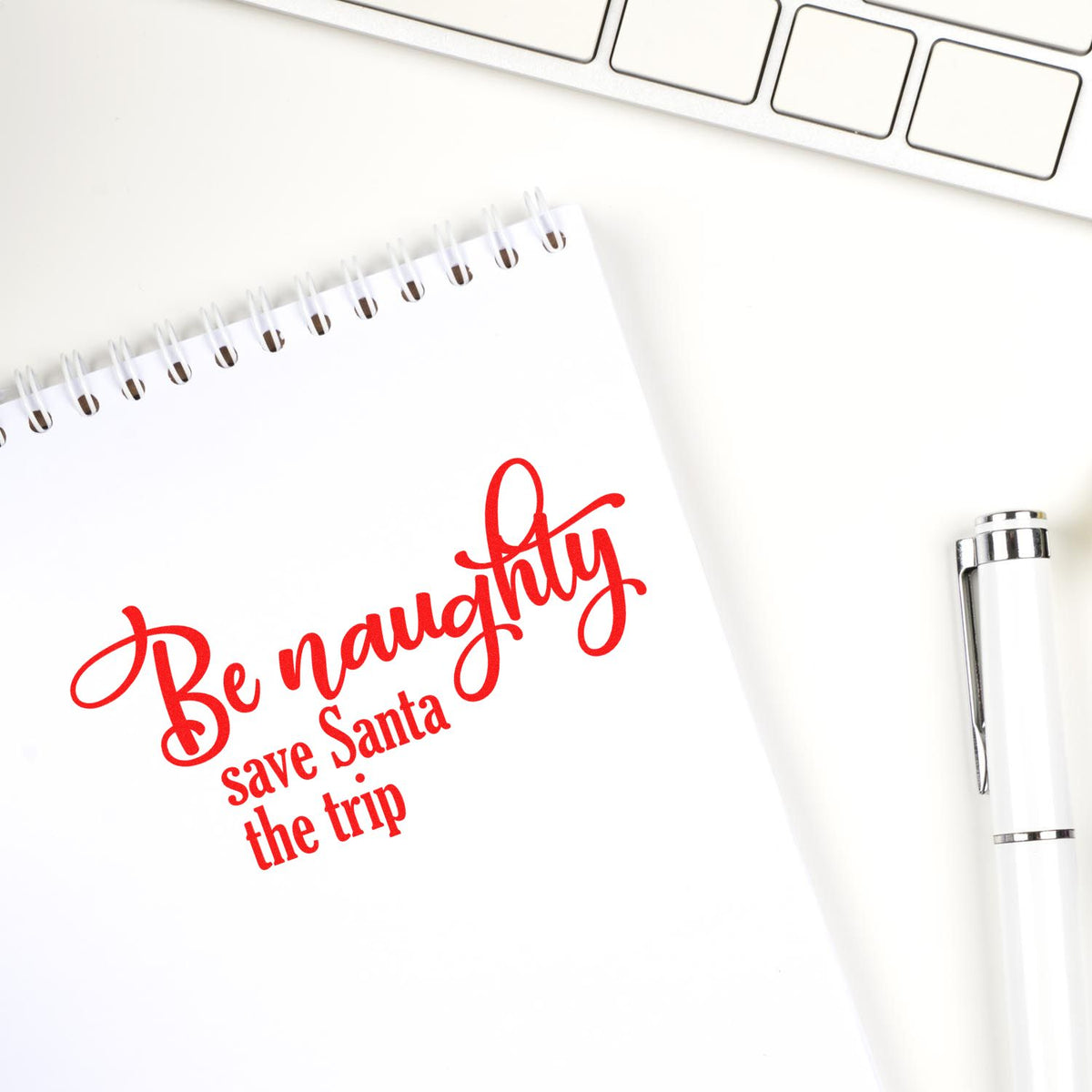 Red holiday rubber stamp reads Be naughty, save Santa the trip on notebook paper with pen and keyboard.