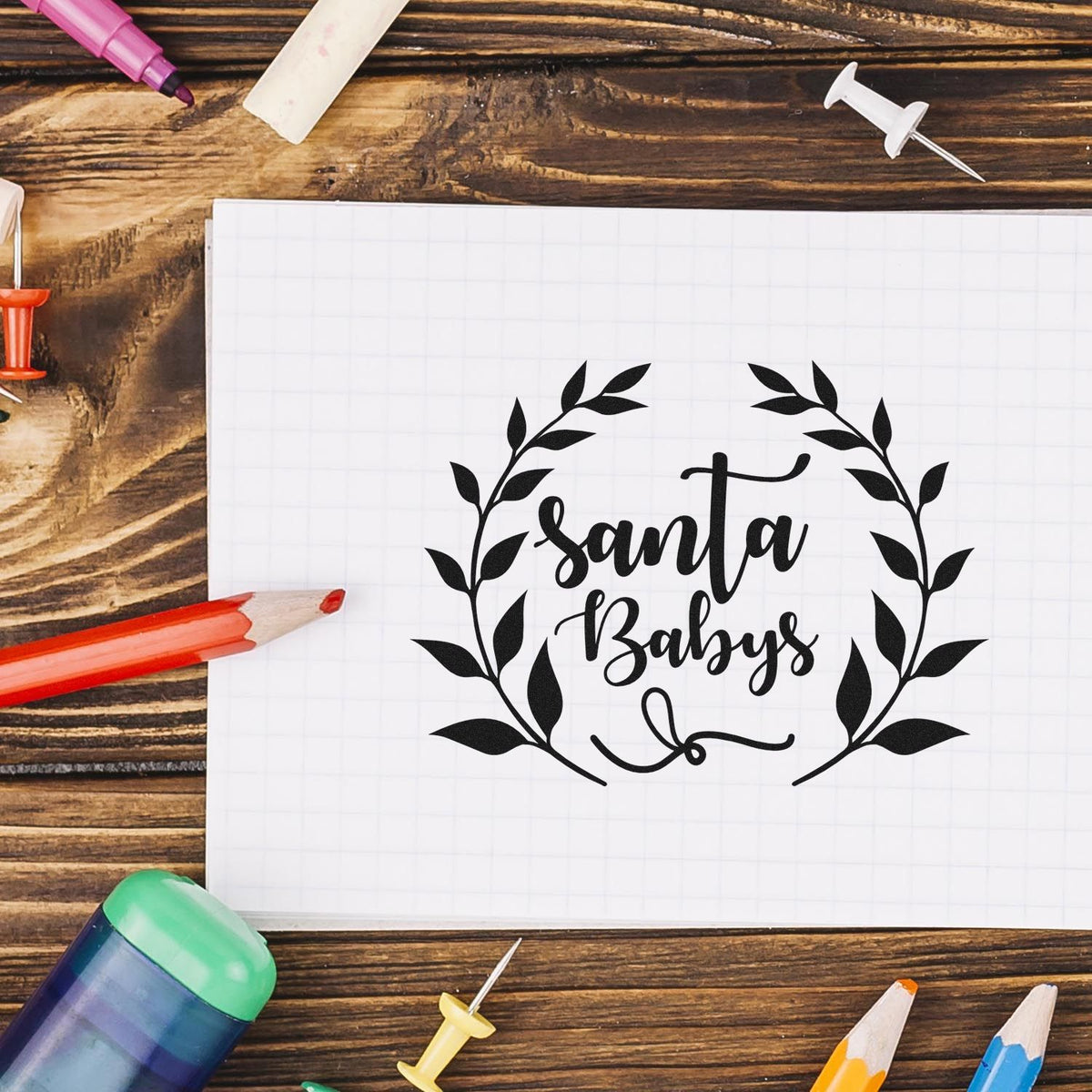 Santa Babys holiday rubber stamp design mockup with leafy wreath on notebook and craft supplies.