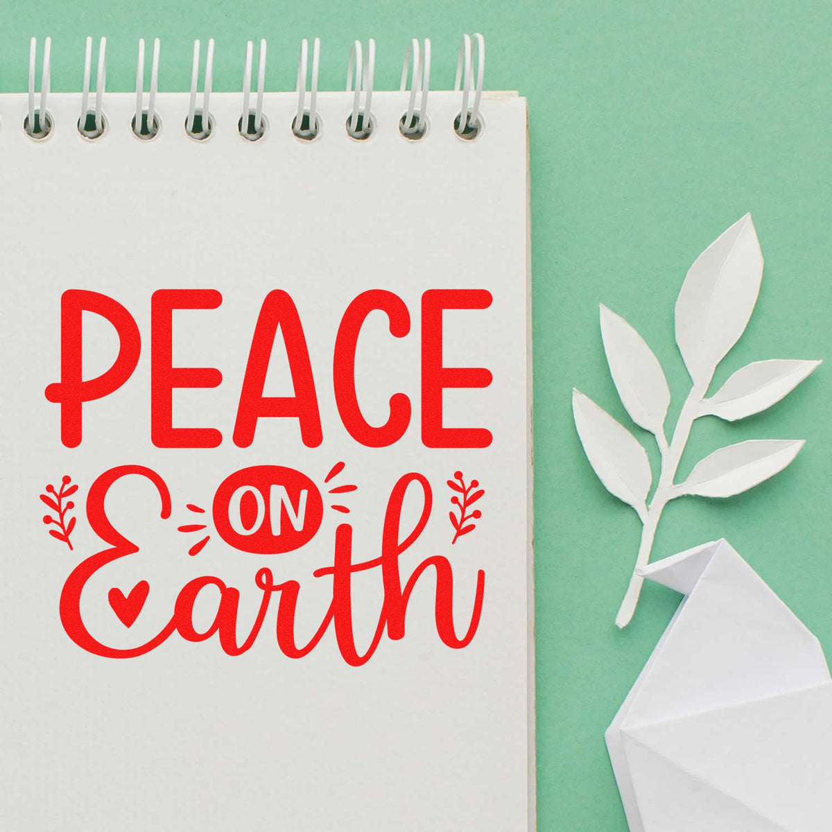Peace on Earth rubber stamp design in red script on notebook, with paper dove and olive branch on mint green background.