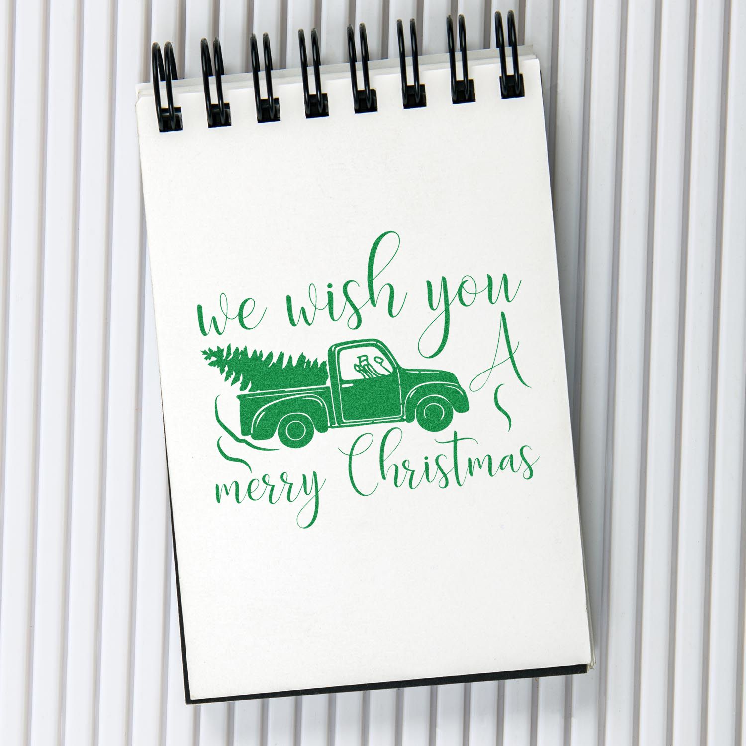 Holiday rubber stamp design featuring a green pickup truck and tree with We wish you a Merry Christmas on a notebook.