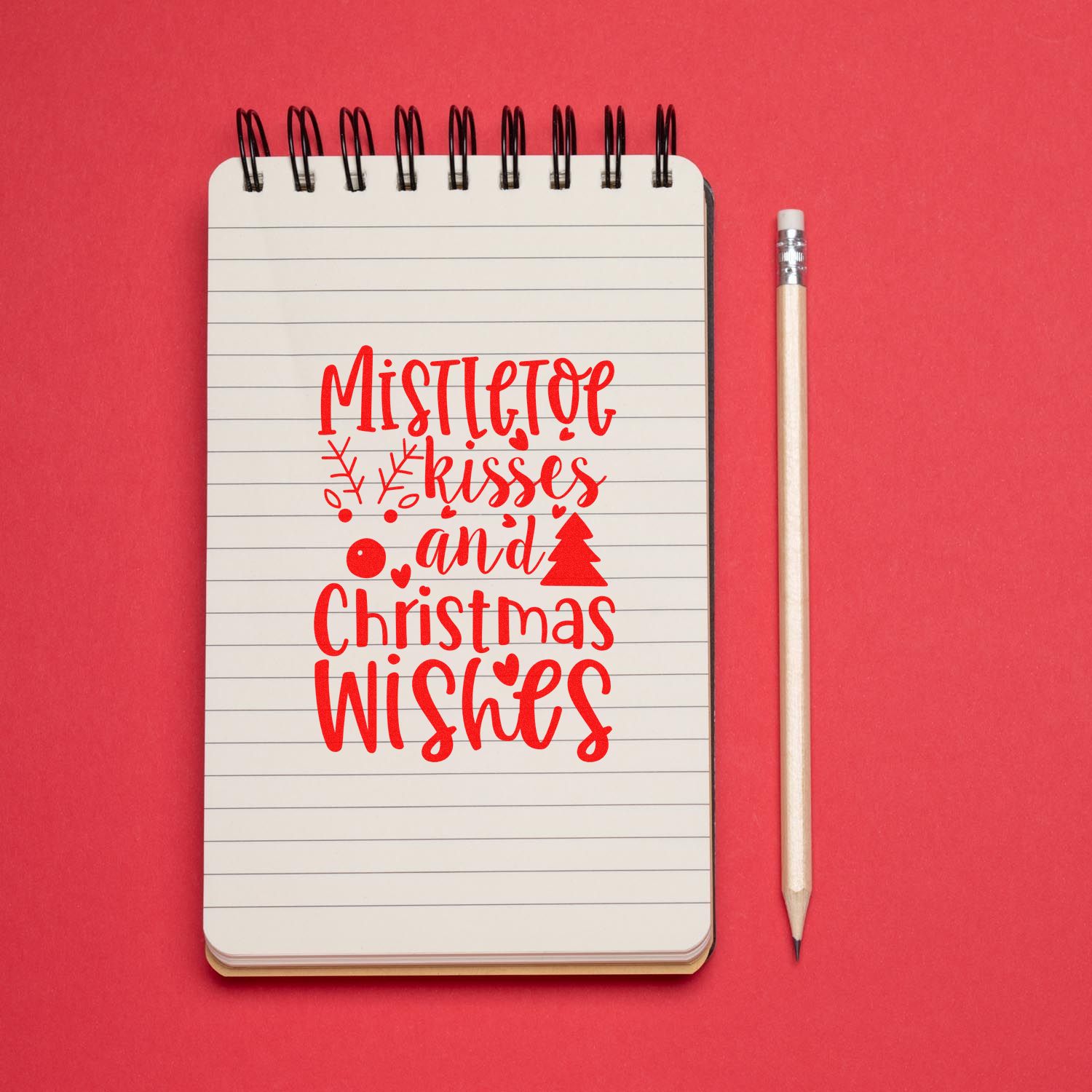 Mistletoe kisses and Christmas wishes rubber stamp design in red on a lined notebook with pencil on a pink background.