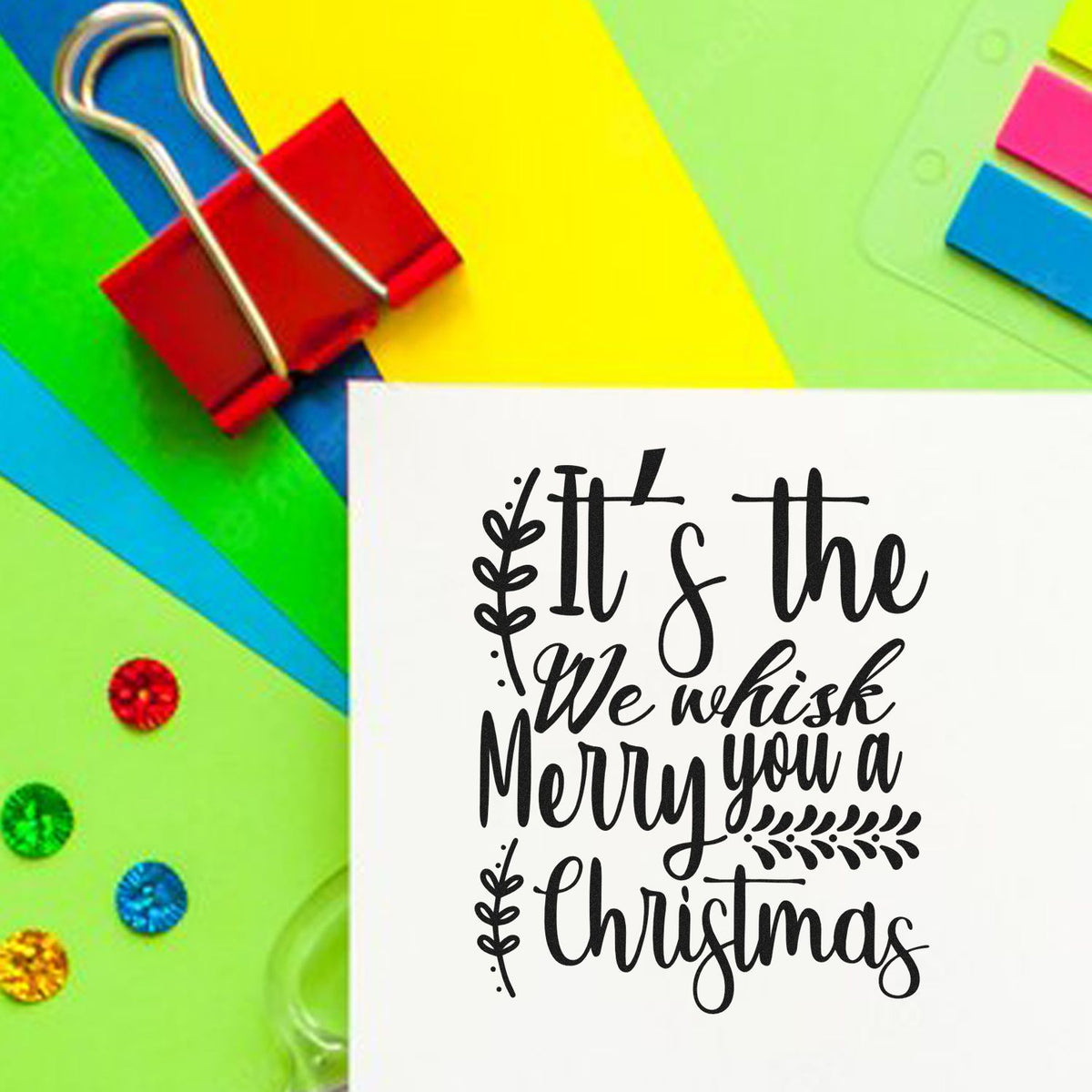Merry Christmas rubber stamp design on handmade card, surrounded by colorful paper clips and craft supplies for holiday card making.