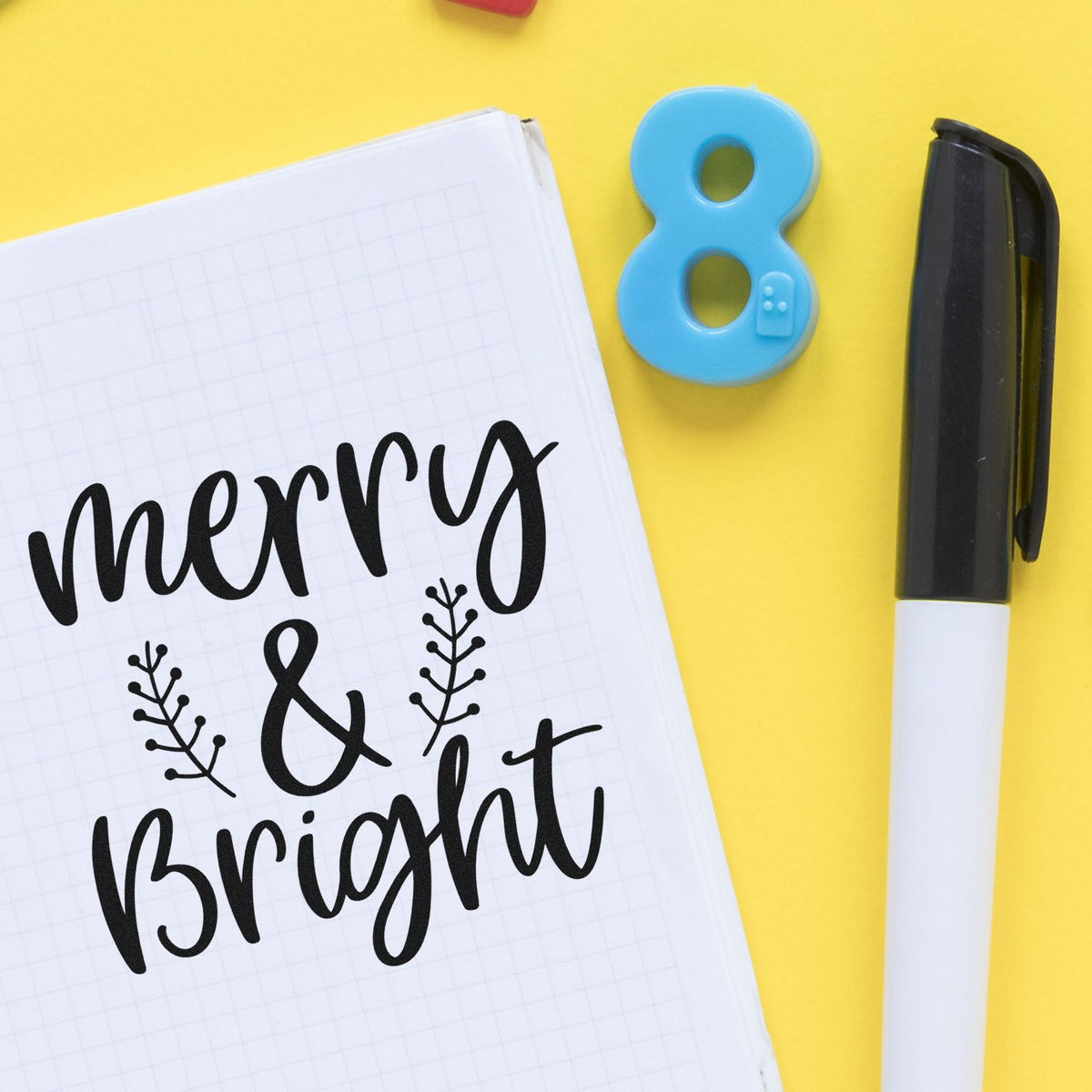 Merry and Bright holiday rubber stamp design in a notebook, with an 8 stamp and marker on a yellow background.