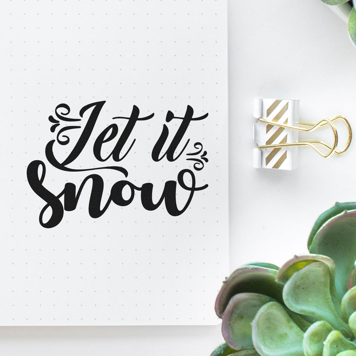 Let It Snow Decorative Script PSI Pre-Inked Stamp imprint stamped in black ink. “Let It Snow” appears in bold script with curled flourishes at both sides.