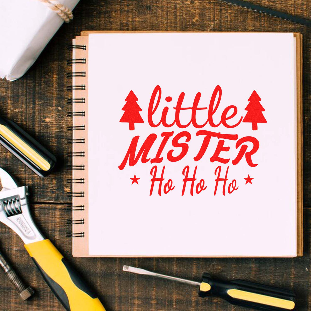 Christmas rubber stamp mockup with Little Mister Ho Ho Ho design on a notebook, surrounded by craft tools.