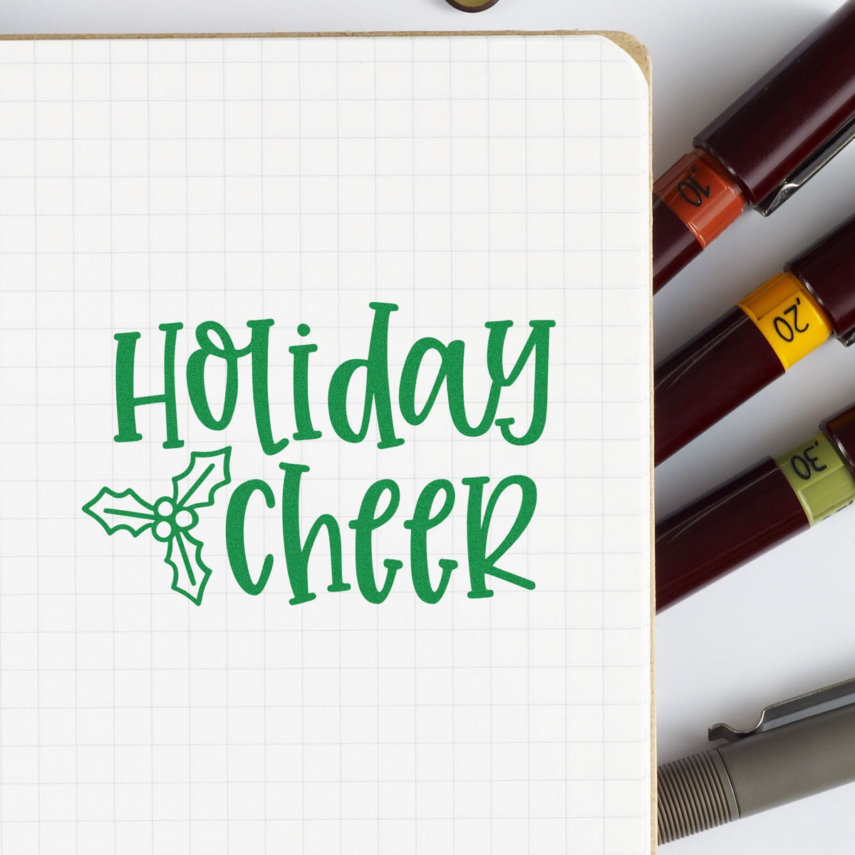 Holiday Cheer rubber stamp in green on a grid notebook, with ink markers for Christmas planner and festive craft projects.