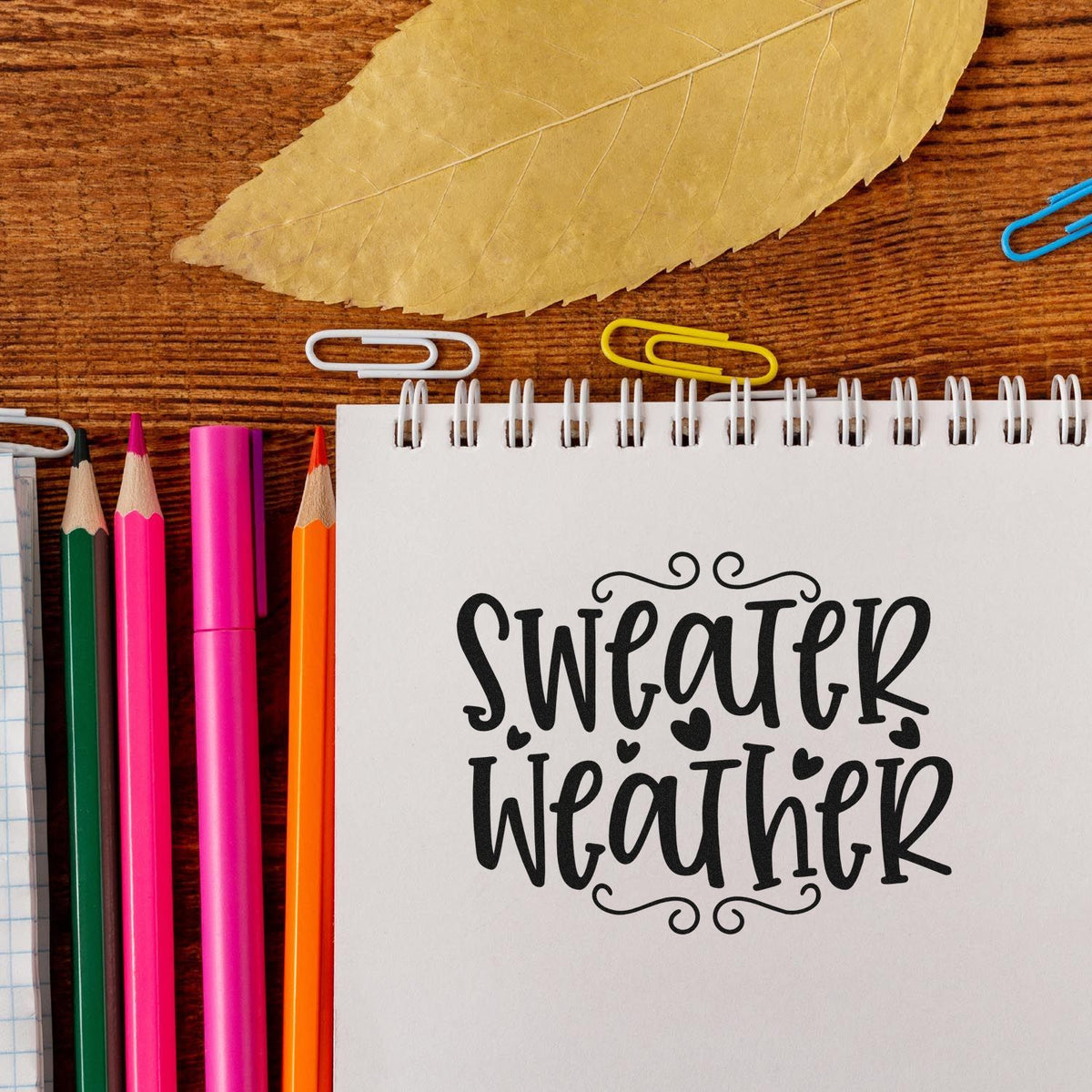 Sweater Weather rubber stamp design on a notebook, styled with pencils and a fall leaf for autumn journaling and crafts.