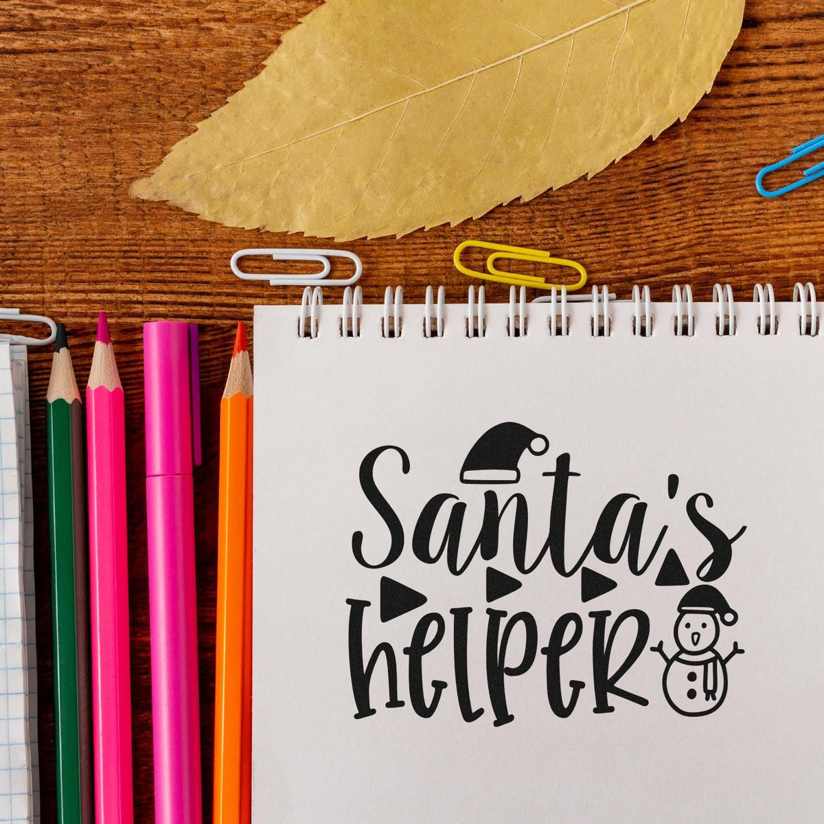 Santa's Helper Christmas rubber stamp design on notebook, surrounded by pencils and paper clips for holiday crafting.