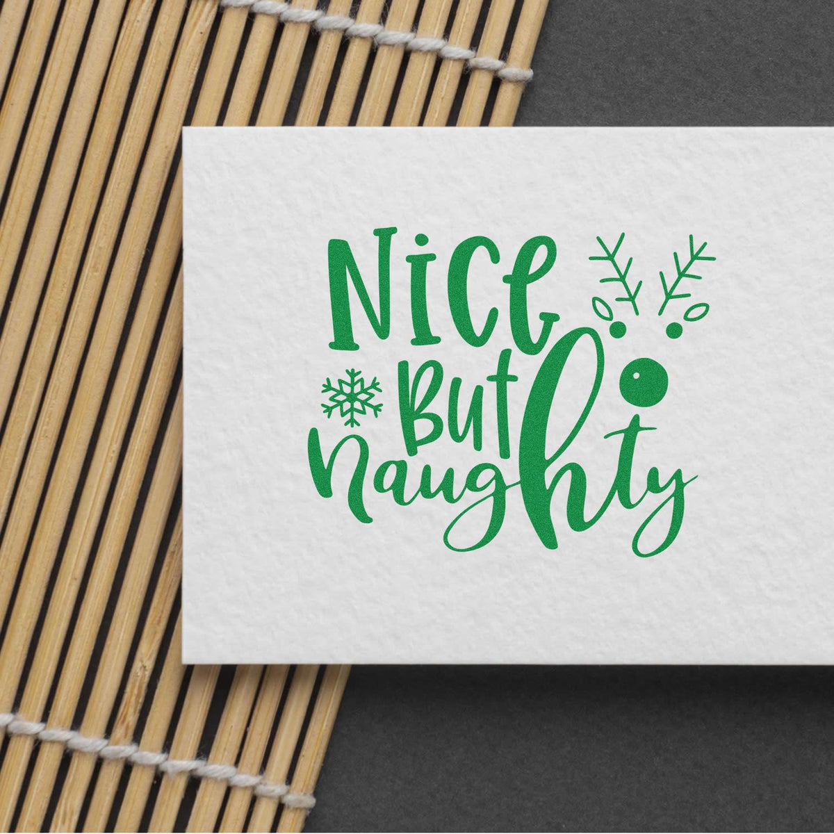 Green Nice but Naughty Christmas rubber stamp impression on white paper with snowflake accents and bamboo mat background.