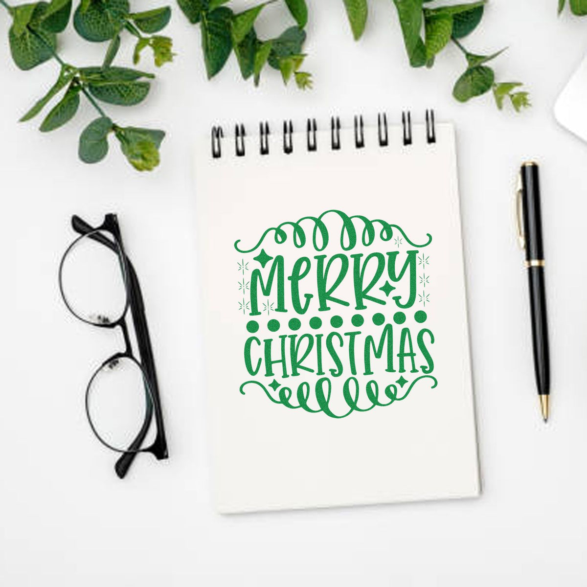 Festive Merry Christmas rubber stamp-style design on notebook with pen, glasses, and greenery for holiday crafting.