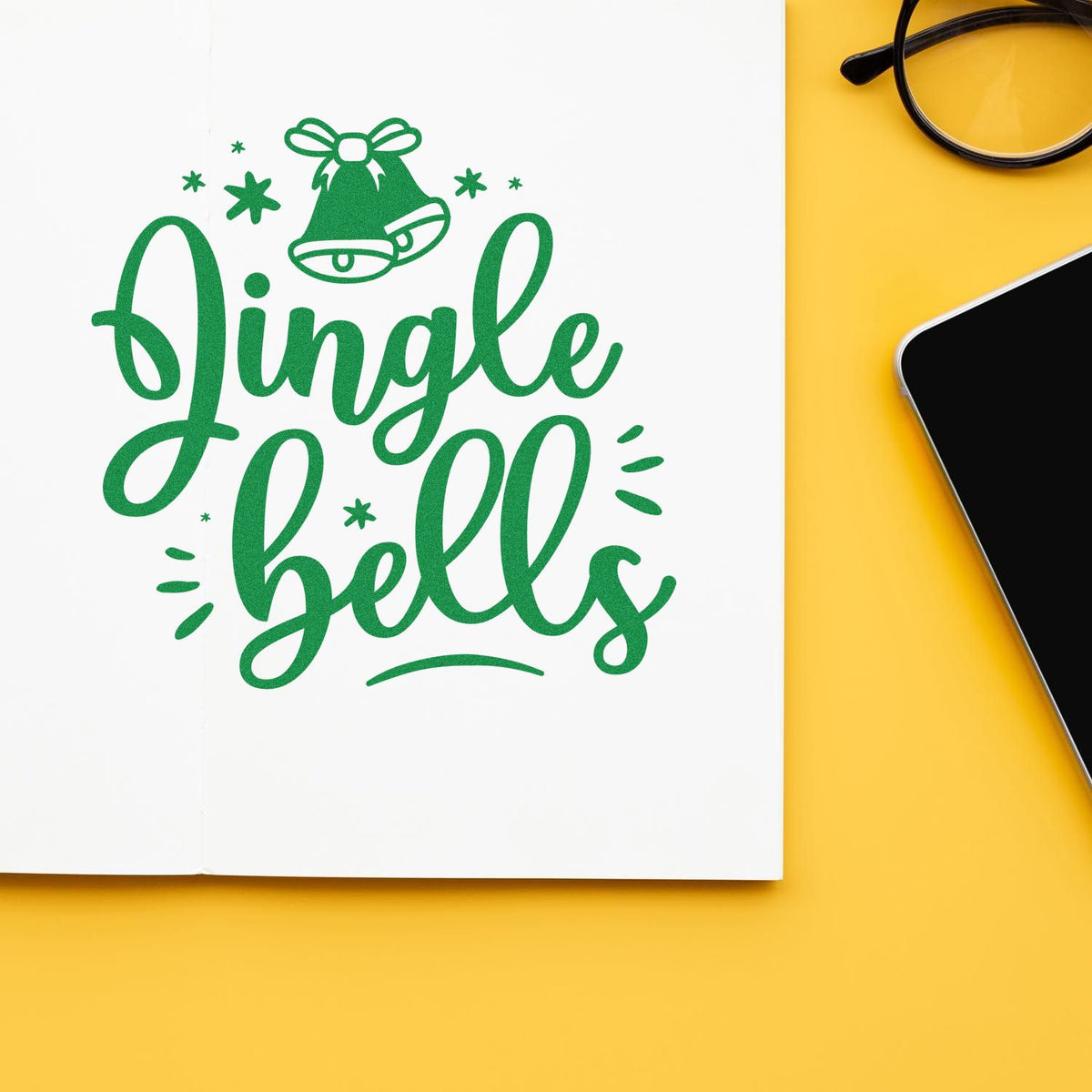 Christmas rubber stamp design with green Jingle Bells lettering and bell icon on a notebook page, ideal for holiday crafts.