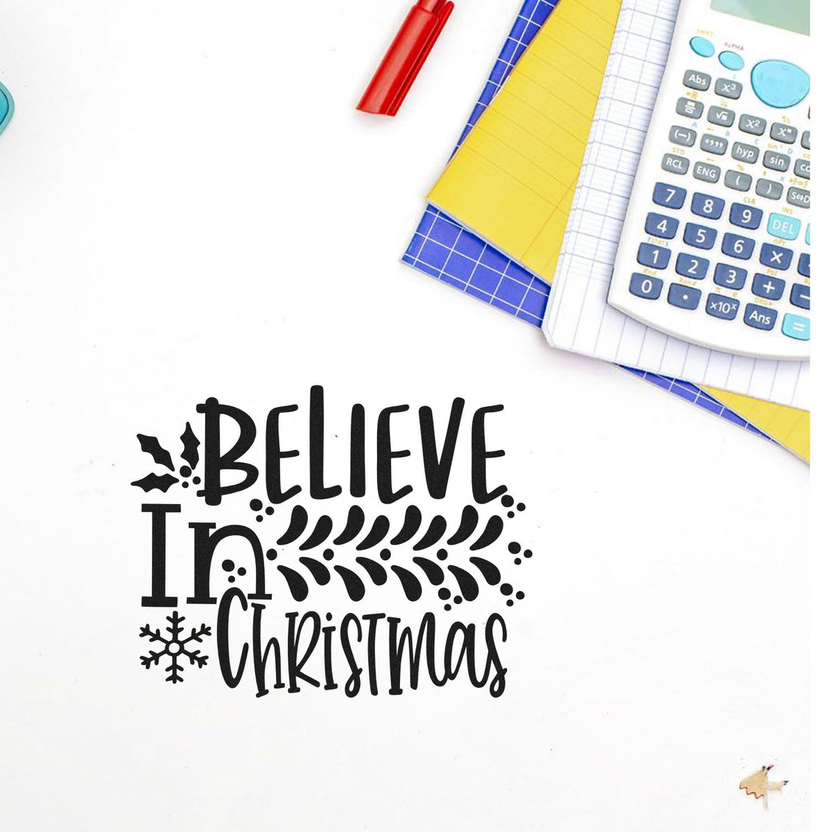 Believe in Christmas rubber stamp design with festive lettering, shown with notebooks and calculator on a clean white background.
