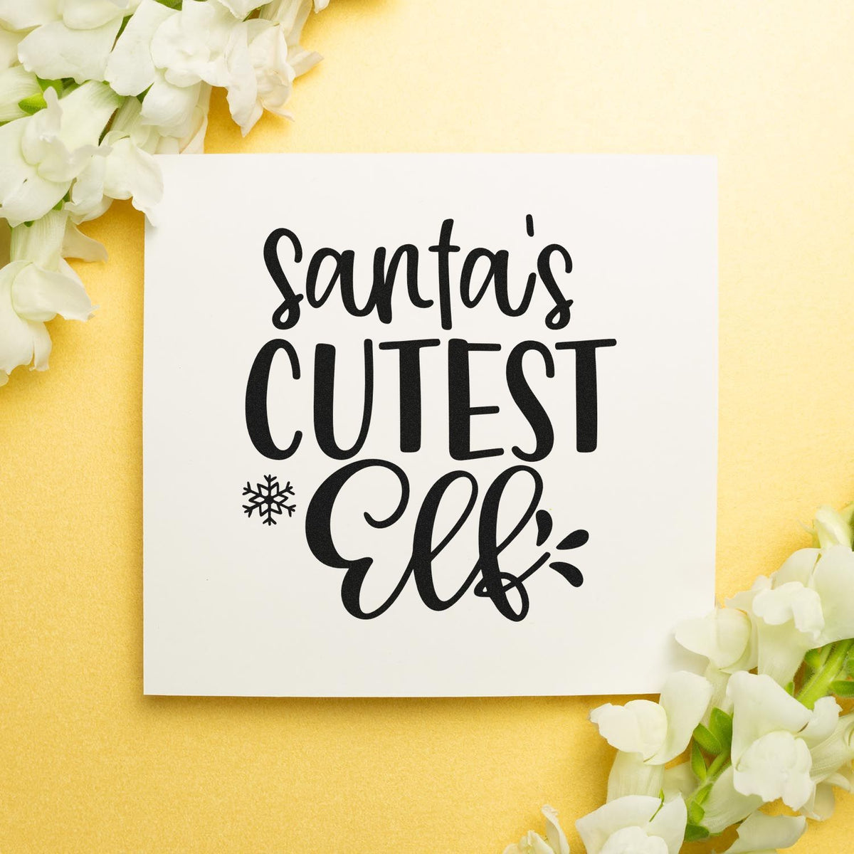 Santa's Cutest Elf Lettering Slim Pre-Inked Stamp imprint stamped in black ink. The words "Santa's cutest elf" appear with a snowflake and small decorative strokes.