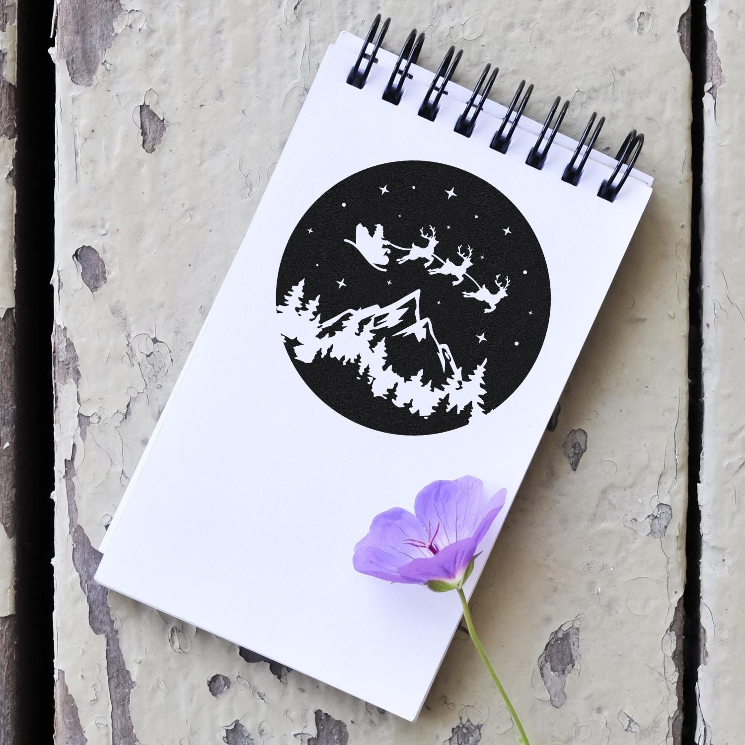 Winter holiday rubber stamp design with Santa sleigh, reindeer, mountains, and pine trees on a notebook page.