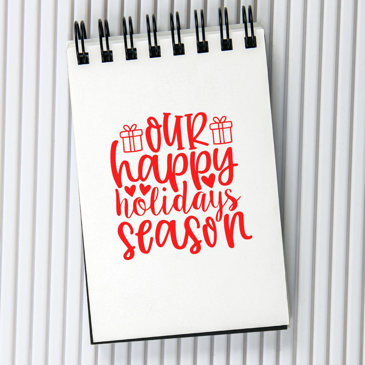 Holiday rubber stamp design with red our happy holidays season lettering, gift icons, and hearts on a spiral notebook.