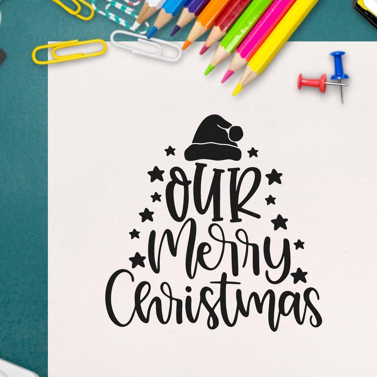 Merry Christmas rubber stamp design on paper, surrounded by colored pencils for festive holiday card making and crafts.