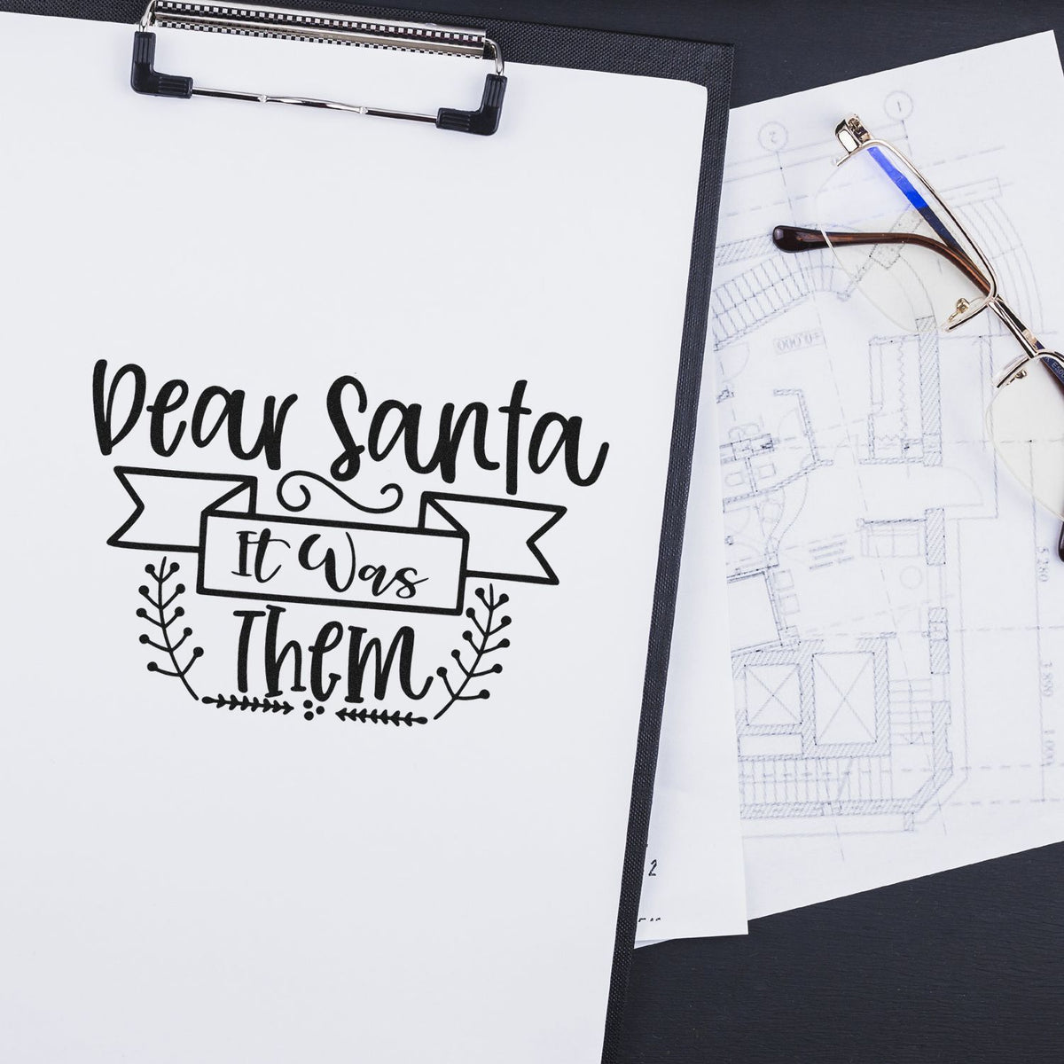 Rubber stamp design reading Dear Santa It Was Them on paper clipboard, shown with eyeglasses and blueprint sketch.
