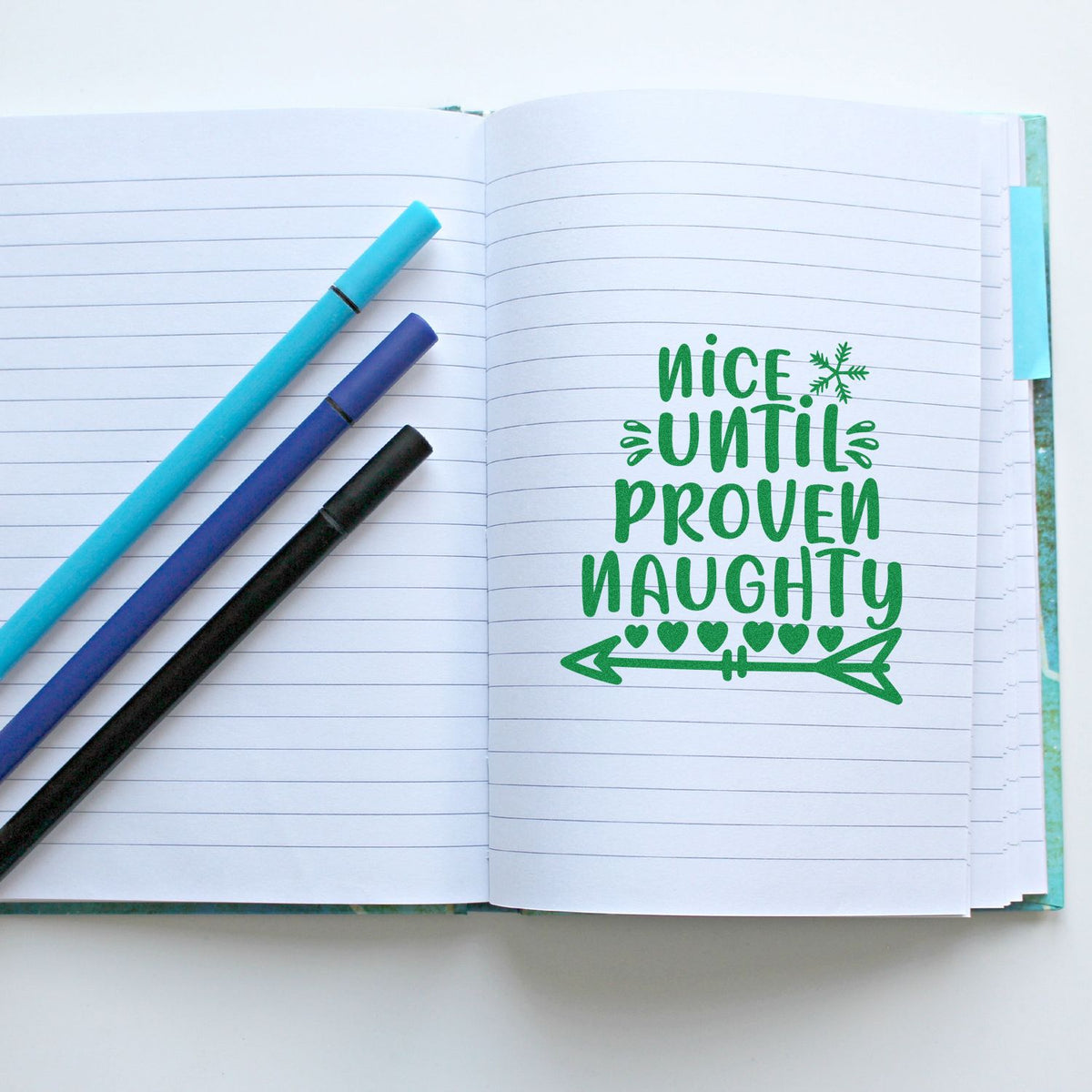 Rubber stamp design on open notebook with green Nice Until Proven Naughty holiday sentiment and arrow graphic.
