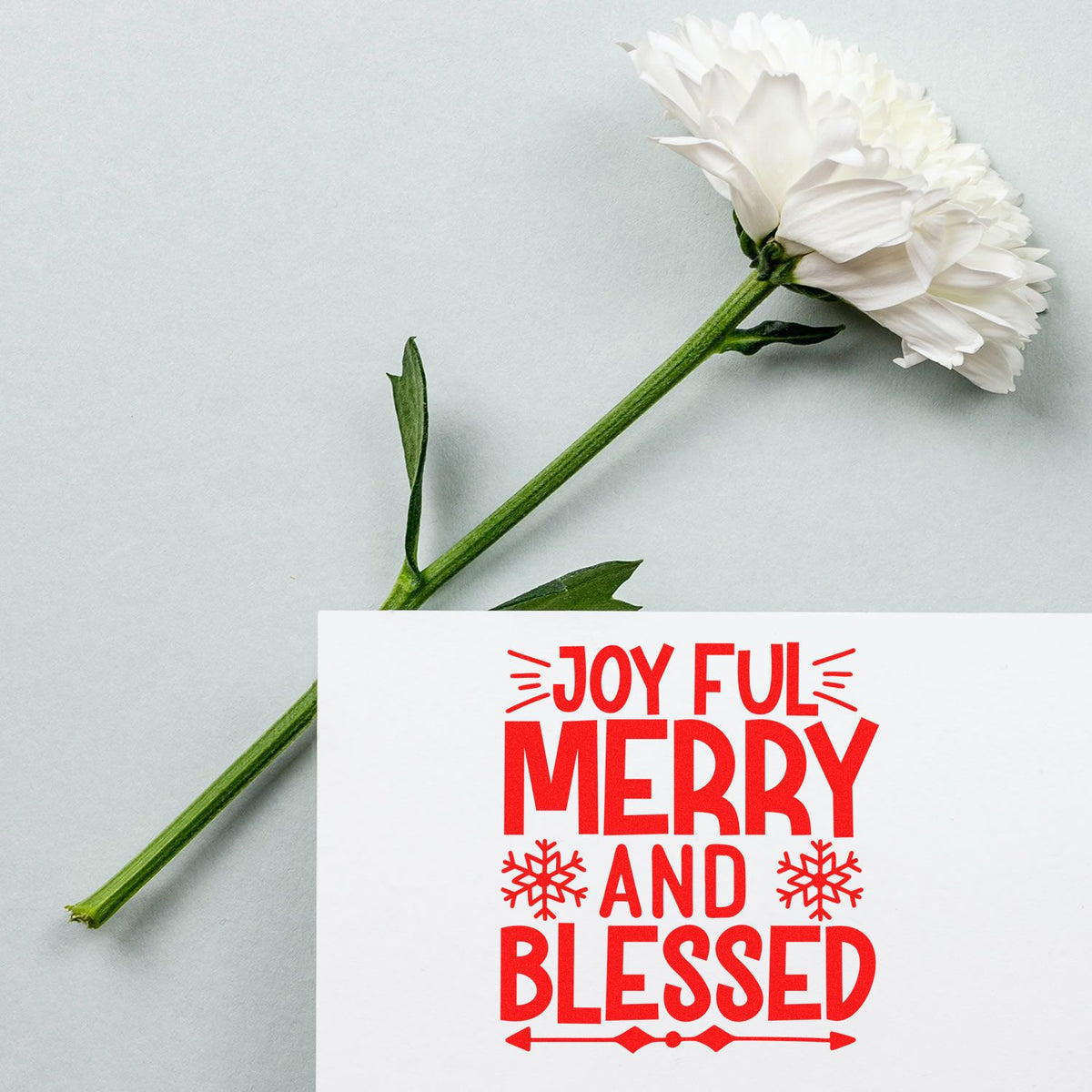 Christmas rubber stamp design reading Joyful Merry and Blessed in red on a white card beside a white flower.