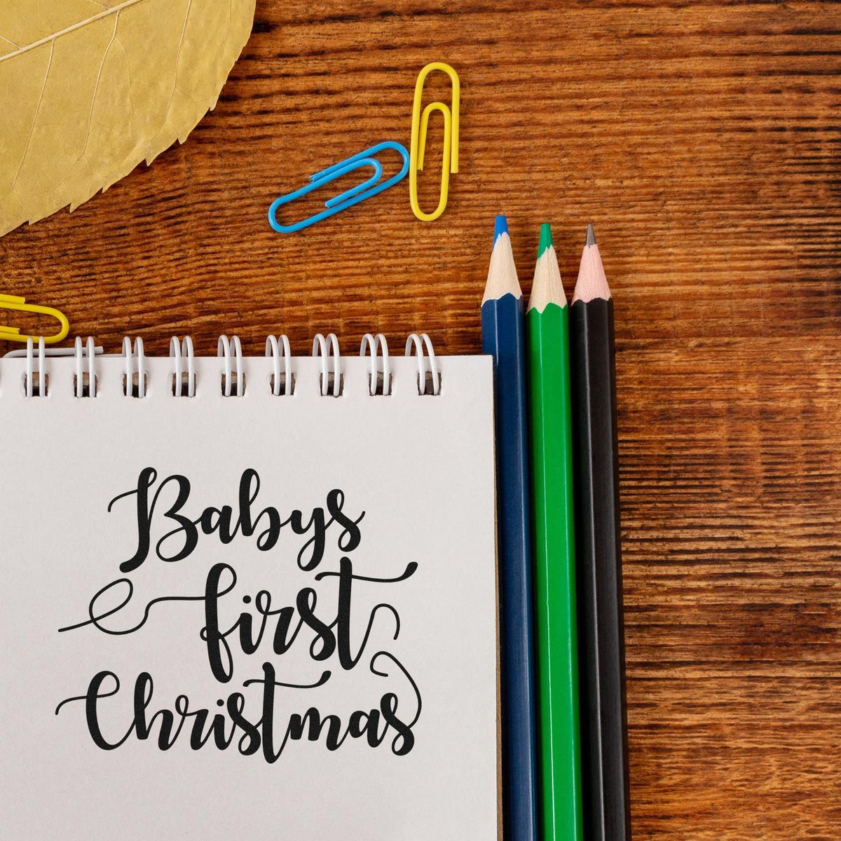 Babys First Christmas rubber stamp design on a notebook, styled with colored pencils and paper clips on a wooden desk