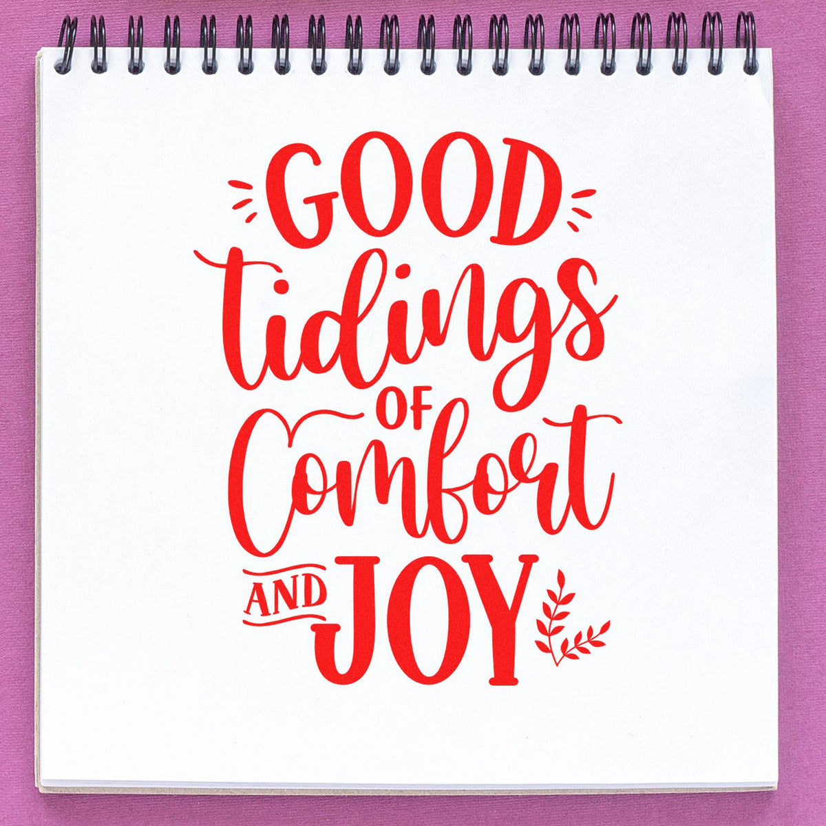Red Christmas rubber stamp design reading Good Tidings of Comfort and Joy on a spiral notebook page.