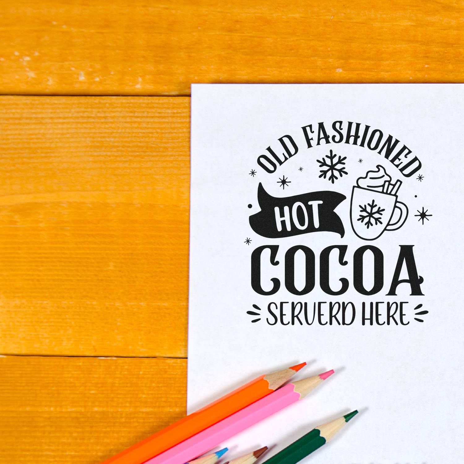 Hot cocoa rubber stamp design with Old Fashioned Hot Cocoa Served Here on white paper, styled with pencils on a wooden table.