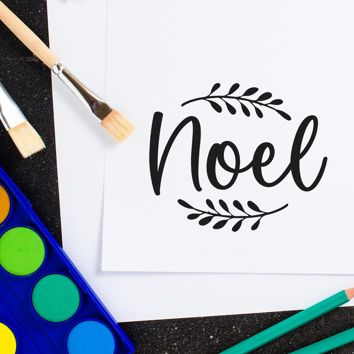 Christmas Noel rubber stamp design with leaf accents on white paper, surrounded by paint brushes and watercolor palette.