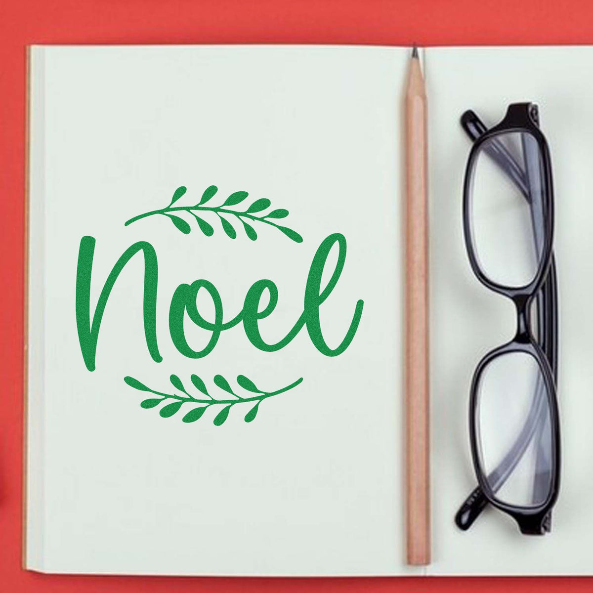 Noel rubber stamp design in green script with leafy accents, styled in a notebook beside a pencil and eyeglasses.
